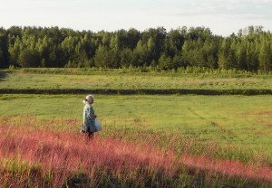 older woman standing in a green field with plants and trees